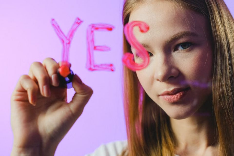 Teenage girl writing the word 'Yes' on glass with marker in studio setting.