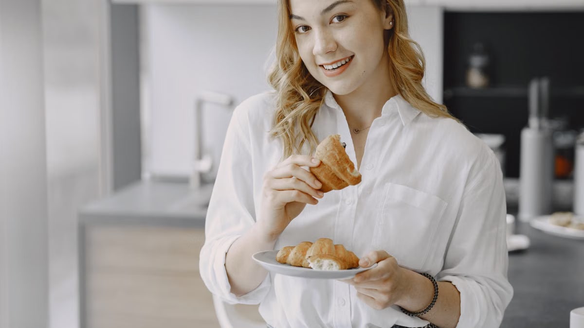 A cheerful woman in a white shirt enjoys fresh croissants in a modern kitchen.